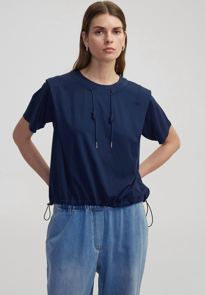 Marineblauwe cropped sweatshirt met korte mouwen, trekkoord in de taille en elastische zoom. Soepele textuur met een casual pasvorm. Gekleed met lichtblauwe jeans.