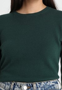 Mörkgrön ribbad stickad tröja med rund halsringning, som har en figurnära siluett och kontrasterande blå denimjeans i midjan.