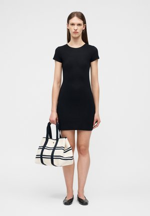 Γυναίκα με μαύρο φόρεμα κοντών μανικιών που κρατά τσάντα χειρός με λευκές και μαύρες ρίγες, φοράει μαύρα φλατ παπούτσια, στέκεται μπροστά σε απλό φόντο.