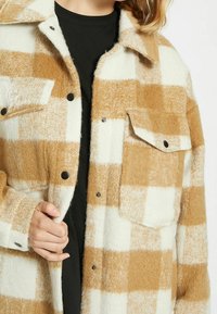 Fleece skjortjacka med ett stort rutmönster i beige och vitt, framknappar, två bröstfickor och en mjuk textur.