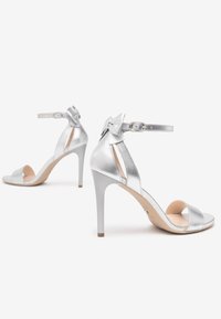 Sandales à talons hauts en argent avec une finition métallique, design à bout ouvert, bride de cheville avec une boucle, et un accent décoratif en forme de nœud.