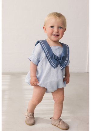 Μικρό παιδί με ξανθά μαλλιά που χαμογελά, φοράει ανοιχτό μπλε φορμάκι με ναυτικό γιακά σε σκούρο μπλε και μπεζ μαλακά παπούτσια, στέκεται σε λείο πάτωμα.