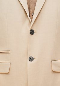 Μπεζ σακάκι με δύο μαύρα κουμπιά κλειστά, γιακά με εγκοπή και τσέπες με καπάκι πάνω σε καφέ πουκάμισο με κουμπιά.