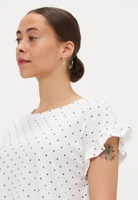 Balta maikutė su juodais taškučiais, raukinėtomis rankovėmis ir laisvu siluetu. Apvalaus kaklo iškirptė. Modelis turi matomą tatuiruotę ant rankos.