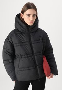 Zwarte pufferjack met een hoge kraag, horizontale stiksels en ritssluiting; gecombineerd met een rode handtas met een polsband.