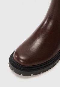 Tmavě hnědá kožená bota s kulatým čtvercovým špičkou a silnou černou lug podrážkou na bílém pozadí.