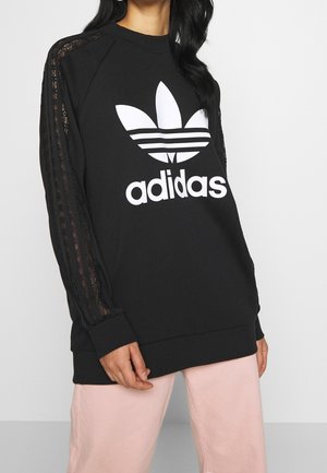 Vrouw draagt een zwarte Adidas-trui met wit logo en kanten strepen op de mouwen, gecombineerd met lichtroze broek.