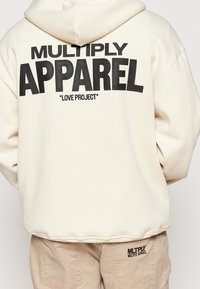 Sudadera beige con un estampado negro que dice "MULTIPLY APPAREL" y "LOVE PROJECT" en la parte de atrás. La tela parece suave y con un corte relajado.