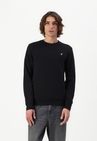 Zwarte sweatshirt gemaakt van zacht materiaal, met lange mouwen en een ronde hals. Een klein gouden logo staat op de linkerborst. Draagt het met grijze jeans.