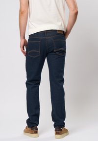 Jeans de mezclilla azul oscuro con un diseño de pierna recta, que cuentan con dos bolsillos traseros y detalles de costura visibles. Combinados con zapatillas de deporte marrones.
