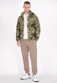 Jachetă de camuflaj în nuanțe de verde, cu glugă, combinată cu pantaloni de trening bej, tapered, și adidași albi cu accente negre.