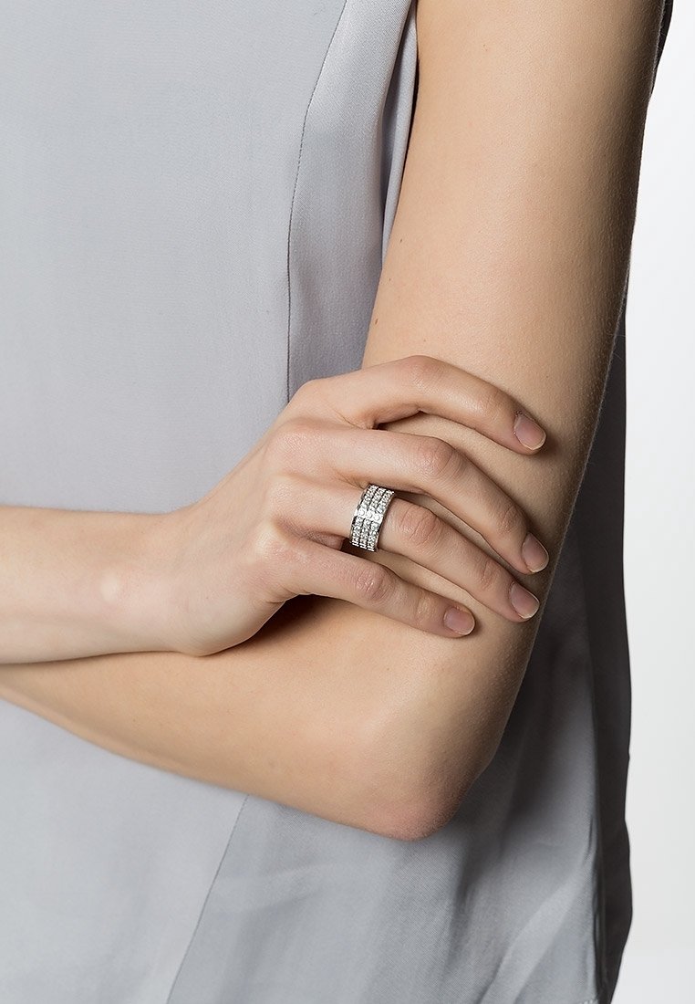 Main avec une large bague en argent ornée de plusieurs rangées de pierres claires, reposant doucement sur le bras opposé portant un haut sans manches gris clair.
