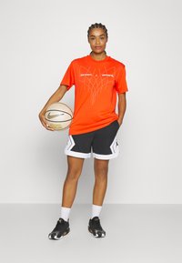 Oranžinė marškinė su grafiniu dizainu, juodi ir balti šortai su šoninėmis detalėmis, baltos sportbačiai, laikantis krepšinio kamuolį.