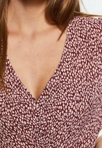 Žena ve vínovém šatu s abstraktním růžovým listovým vzorem a výstřihem do V, který odhaluje holá ramena a horní část hrudníku.