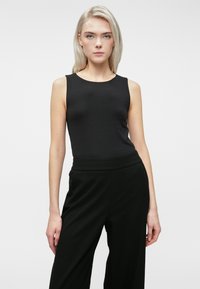 Mlada žena z blond lasmi, oblečena v črno brezrokavno majico in črne hlače visoko pasu, stoji pred enostavno svetlo ozadje.