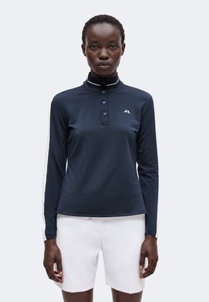 Frau trägt ein marineblaues Langarm-Poloshirt mit weißen Streifen an den Ärmeln und weiße Shorts, steht vor einem schlichten hellen Hintergrund.