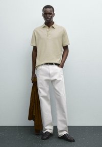 Beige polo tričko, volné bílé kalhoty a černé sandály. Drží hnědý kabát. Minimalistické pozadí a neutrální tóny.