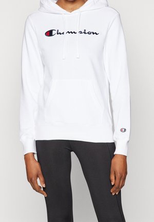 Person iført en hvid Champion hættetrøje med logo på bryst og ærme, kombineret med sorte leggings, stående mod en ensfarvet baggrund.