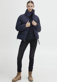 Námořnická puffer bunda s vysokým límcem, spárovaná s tmavým pleteným svetrem, úzkými černými džíny a hnědými kotníkovými botami se žlutými tkaničkami.