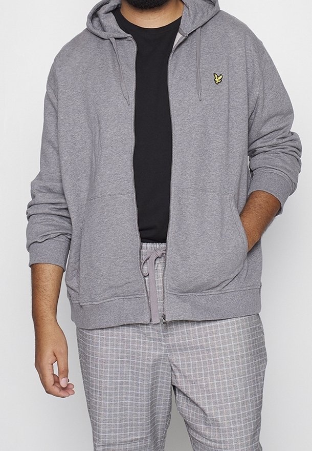 Homme portant un sweat à capuche gris zippé avec un logo d'oiseau jaune, un t-shirt noir et un pantalon à carreaux gris avec une taille à cordon.