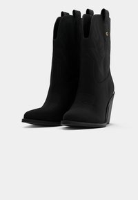 Botines de ante negros hasta el tobillo con punta afilada, costuras decorativas, lengüetas en el tirador y tacón de cuña. Presentan un detalle de marca dorada en el lateral.