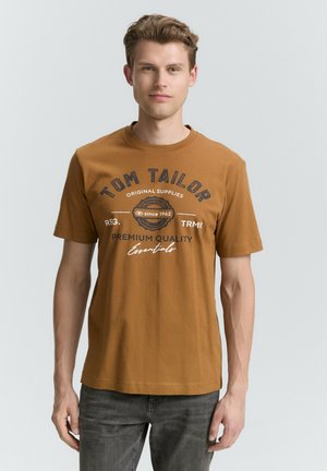 Bruine katoen t-shirt met ronde hals, korte mouwen en een geprinte logo in donkerblauw, met tekst en een circulair ontwerp. Gemaakt met donkere spijkerbroeken.