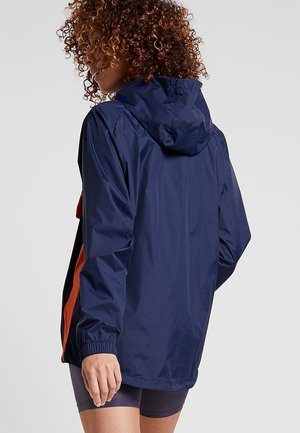 Krøllet person iført en mørkeblå hættetrøje med orange sidepaneler, set bagfra mod en ensfarvet lys baggrund.