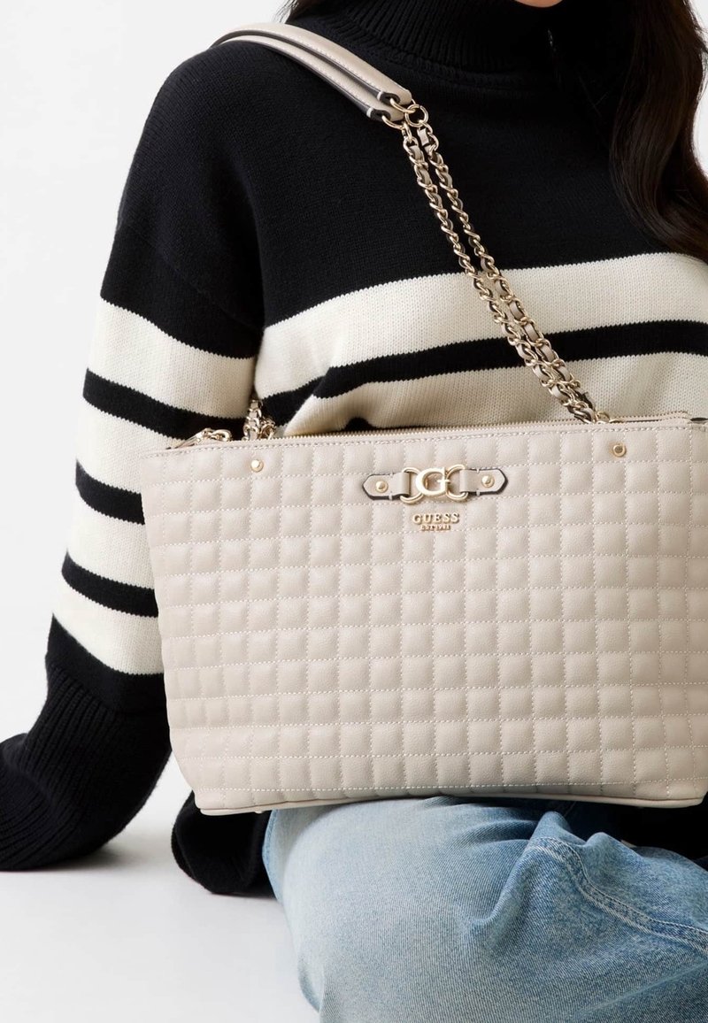 Personne portant un pull rayé noir et blanc et un jean bleu, tenant un sac à main matelassé beige avec une bandoulière chaîne dorée et un logo sur le devant.