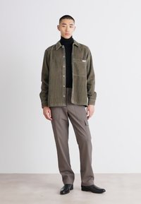 Olive green corduroy shirt jacket with a pocket, worn over a black turtleneck. Brown cargo pants and black shoes complete the outfit.