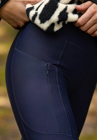Tummansiniset legginssit, joissa on sileä pinta, reisissä vetoketjullinen tasku ja vaaleat ompeleet saumoissa.