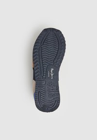 Suela de zapato de goma oscura con un patrón de relieve prominente. Presenta una mezcla de materiales en azul marino, beige y azul. Logo visible en el lateral.