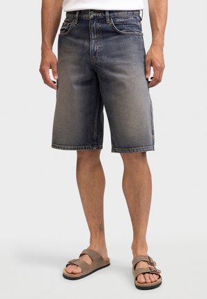 Muž v opraných čiernych riflových šortkách a hnedom dvojpásikovom sandále, stojaci na jednoduchom svetlo sivom pozadí.