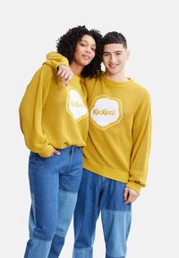 Des pulls en tricot jaunes avec un logo de nuage blanc, portés avec un jean en denim bleu clair présentant un patchwork. Coiffures bouclées et lisses.