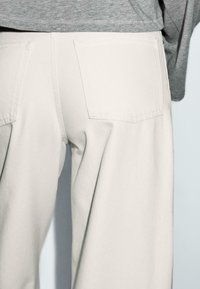 Pantaloni larghi beige chiaro con due tasche posteriori e un tessuto liscio e strutturato. Abbinati a una maglietta grigia a maniche corte.