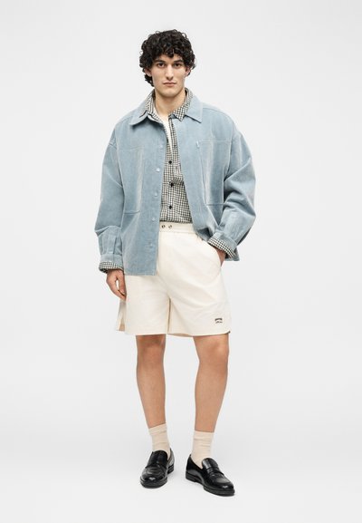 Jeune homme portant une veste surdimensionnée bleu clair, une chemise à carreaux, un short crème, des chaussettes beiges et des mocassins en cuir noir, debout devant un fond uni.