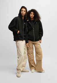 Felpe con cappuccio nere con zip, vestibilità ampia, indossate sopra magliette nere. Una con pantaloni cargo crema, l'altra con pantaloni cargo beige. Calzature di colore chiaro.