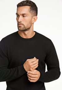 Czarny sweter z długim rękawem i okrągłym dekoltem, wykonany z gładkiego, teksturowanego materiału. Charakteryzuje się prostym wzornictwem, dopasowanymi mankietami oraz subtelnymi detalami w przeszyciach.