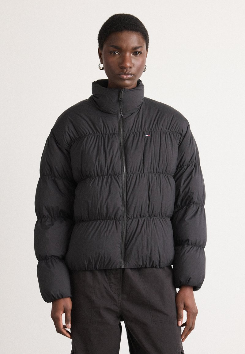 Svart boblejakke med høy krage, horisontale baffel, og glidelås foran. Har en liten logo på brystet, med matt finish og glatt tekstur.