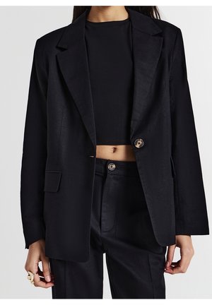 Kobieta ubrana w czarny krótki top pod oversize'ową czarną marynarką oraz pasujące, czarne, wysokie spodnie, odsłaniające brzuch i z guzikami.