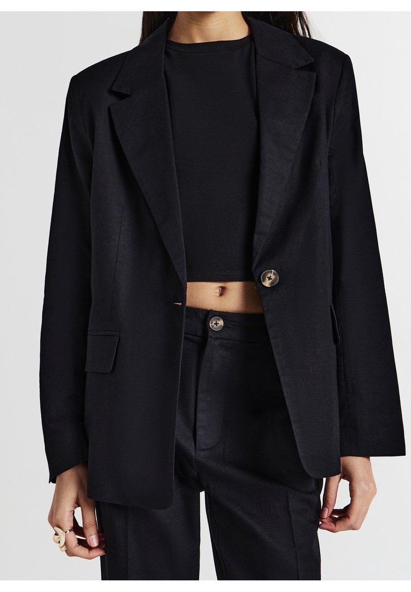 Femme portant un top court noir sous un blazer noir oversize assorti à un pantalon taille haute noir, dévoilant le ventre et des détails de boutons.