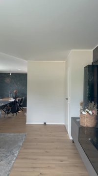 Interni moderni con pavimenti in legno chiaro, pareti bianche, un armadio scuro con un cesto intrecciato e una vista su una zona pranzo con sedie nere.