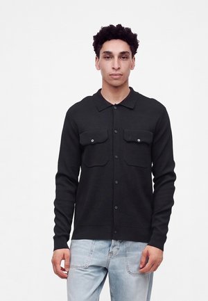 Homme aux cheveux courts et bouclés porte une chemise noire boutonnée avec deux poches avant et un jean bleu clair, debout devant un fond blanc uni.