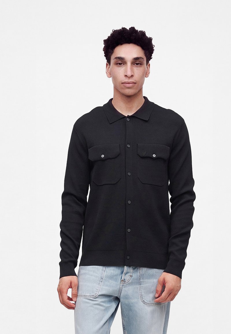 Homme aux cheveux courts et bouclés porte une chemise noire boutonnée avec deux poches avant et un jean bleu clair, debout devant un fond blanc uni.