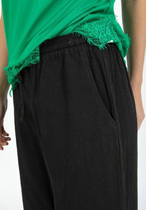 Detailný záber osoby nosiacej voľné čierne nohavice s elastickým pásom a zeleným topom s čipkovaným lemom, čiastočne zastrčeným dovnútra.