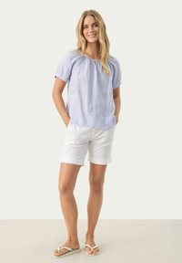 Hellblaue Leinenbluse mit kurzen Puffärmeln, kombiniert mit weißen, hochgekrempelten Shorts und weißen Sandalen. Einfacher, lockerer Schnitt und lässiger Stil.