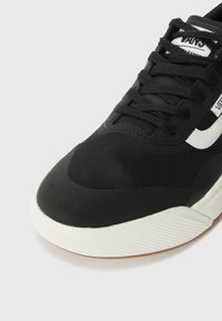 Svart sneaker med overdel i syntetisk tekstil, hvite logo detaljer, rund tå og en teksturert gummisåle.
