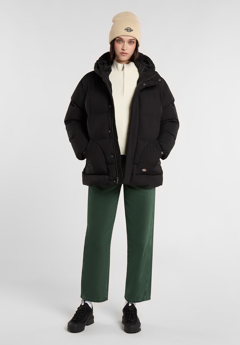 Chaqueta de plumas negra con capucha, cremallera frontal y bolsillos laterales. Llevada sobre un suéter color crema, combinada con pantalones de pierna ancha verde y zapatillas negras.