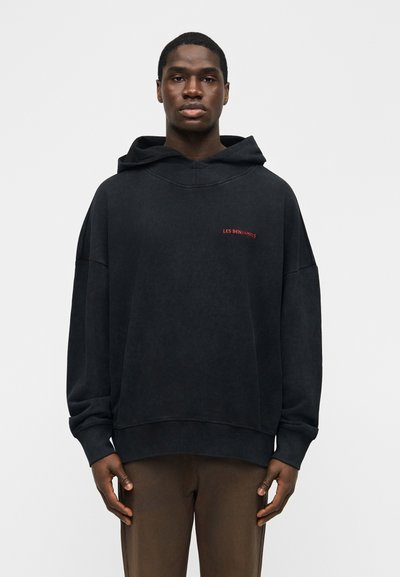 Jeune homme portant un sweat à capuche noir oversize avec le texte rouge "LES BENJAMINS" et un pantalon marron, debout devant un fond blanc uni.