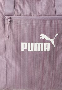 Violetti raidallinen olkalaukku, joka on valmistettu teksturoidusta kankaasta. Siinä on etupuolella vetoketjullinen tasku ja suuri valkoinen PUMA-logo. Yksinkertainen, toiminnallinen muotoilu.