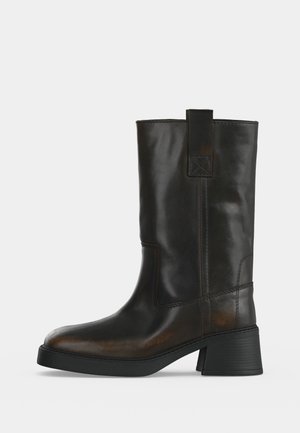 Botte en cuir noire mi-mollet avec un passant, un bout rond et un talon bloc sur un fond clair uni.